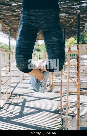Vertical image of an athletic young woman doing pull-ups outdoors in the morning, close-up. Female legs in blue sneakers. Stock Photo