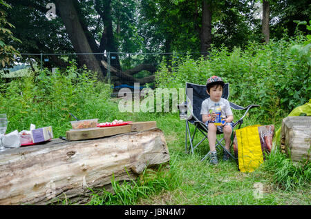 A young bot sits in a folding chair on a camp site and snjoys a drink.