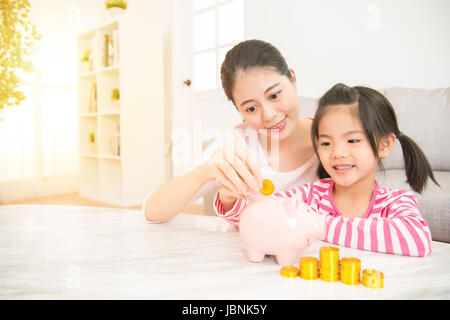 happy asian chinese mother and daughter putting coins into piggy bank in the living room at home. family activity concept. Stock Photo