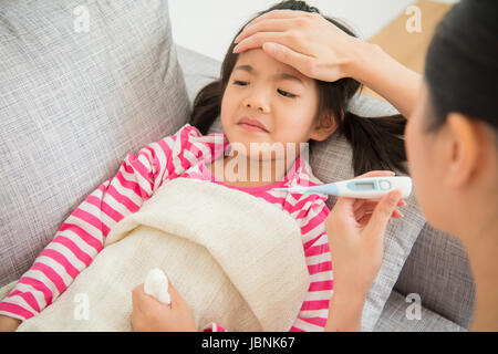 Cute little asian girl is ill lying on sofa bed mother checking a thermometer and touching daughter's forehead in the living room at home. family acti Stock Photo