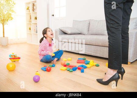 gloat little asian kid mess up all over of living room sitting on wooden floor and hard working business mother back from work feel shocked and angry. Stock Photo