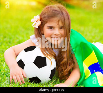 Closeup portrait of cute little football fan lying down on fresh green grass with ball and big Brazil flag, happy supporter of Brazilian football team Stock Photo