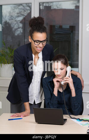 Eine junge Büroangestellte sitzt am Schreibtisch vor einem Notebook und telefoniert. Eine weitere lächelnde Frau steht neben ihr. Stock Photo