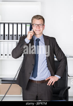 Junger Geschäftsmann mit Brille steht am Schreibtisch im Büro und telefoniert. Im Hintergrund befindet sich ein Regal. Der Mann schaut zur Kamera und lächelt. Stock Photo