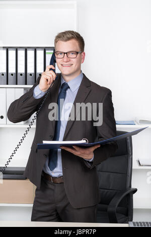 Junger Geschäftsmann mit Brille steht am Schreibtisch im Büro und telefoniert. In der einen Hand hält er einen Aktenordner. Im Hintergrund befindet sich ein Regal. Der Mann schaut zur Kamera und lächelt. Stock Photo