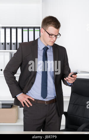 Junger Geschäftsmann mit Brille steht im Büro vor einem Regal und schaut konzentriert auf sein Smartphone. Stock Photo