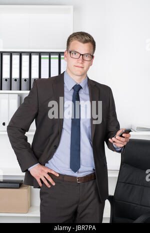 Junger Geschäftsmann mit Brille steht im Büro vor einem Regal und hält dabei in der Hand ein Smartphone. Der Mann schaut zur Kamera. Stock Photo