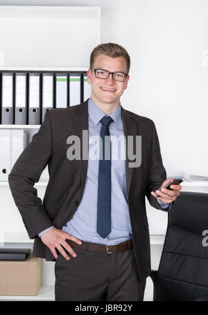 Junger Geschäftsmann mit Brille steht im Büro vor einem Regal und hält dabei in der Hand ein Smartphone. Der Mann schaut zur Kamera und lächelt. Stock Photo