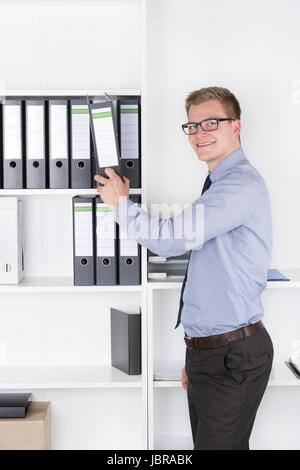 Junger Geschäftsmann mit Brille steht vor einem Regal im Büro und zieht einen Aktenorder aus dem Regal heraus. Der Mann schaut zur Kamera und lächelt. Stock Photo