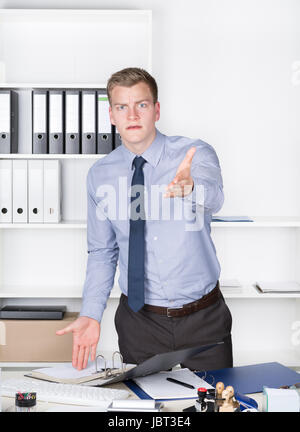 Junger wütender Geschäftsmann steht vor einem Regal im Büro und streckt einem Arm aus in Richtung Kamera. Der Mann schaut zur Kamera.