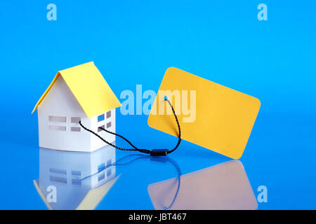 Paper house attached to yellow blank price tag on blue background Stock Photo