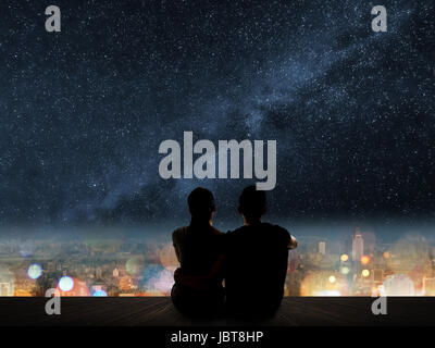 Silhouette of young Asian couple sit on wooden ground above the city under stars. Stock Photo