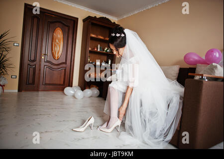 Pretty bride putting on elegant white wedding shoes in her room. Stock Photo