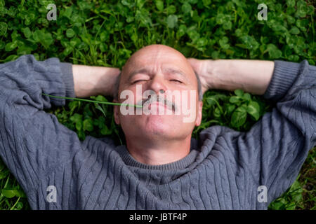 Mature Happy Man Lying On Green Grass. He holds onion leaf in mouth and closes eyes. Resting on nature Stock Photo