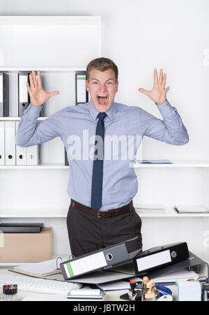Junger wütender Geschäftsmann steht vor einem Berg von Akten auf seinem Schreibtisch im Büro und schreit dabei. Im Hintergrund befindet sich ein Regal. Der Mann schaut zur Kamera.