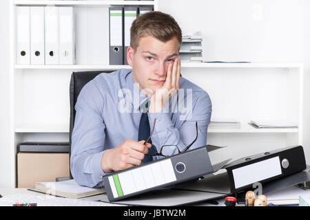 Junger nachdenklicher Geschäftsmann sitzt vor einem Berg von Akten auf seinem Schreibtisch im Büro. Im Hintergrund befindet sich ein Regal. Der Mann schaut zur Kamera. Stock Photo