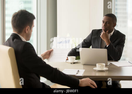 Two multiracial businessmen on corporate meeting in suits discussing reading financial report with rising graphs, showing results of successful teamwo Stock Photo