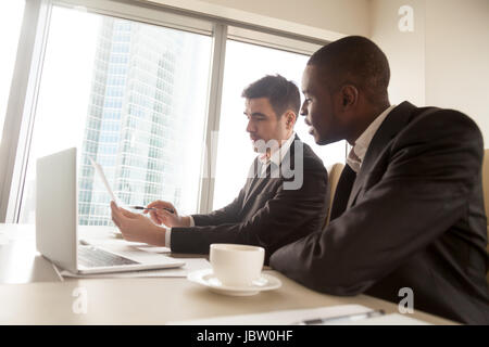 Side view of two multiracial business partners in formal wear discussing document on meeting sitting at desk, black and white businessmen studying con Stock Photo
