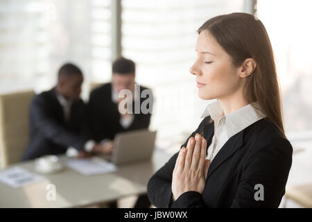 Young beautiful businesswoman enjoys meditating standing in office with eyes closed, put hands together as in prayer, reduce stress, relief to keep ca Stock Photo