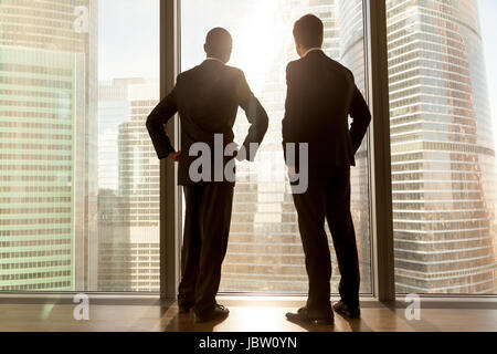 Rear view of african and caucasian businessmen wearing suits standing at full length large window enjoying great big city scenery, contemplative thoug Stock Photo