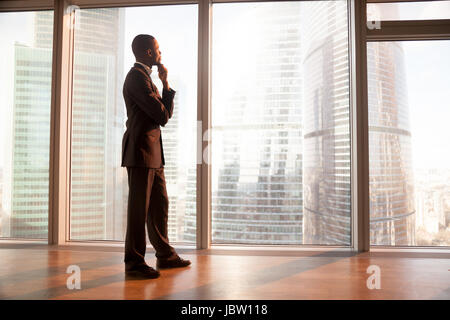 Young afro american contemplative businessman stands in office with hand on chin, looks through big window at the city, african entrepreneur enjoys su Stock Photo