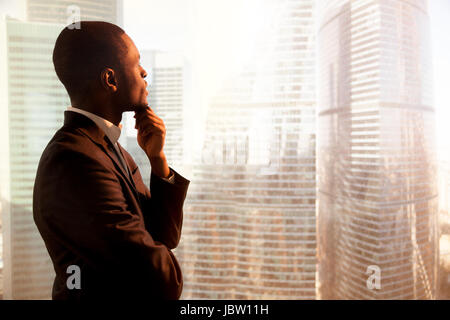 Young african-american thoughtful businessman wearing suit holding hand on chin, looking out of big office window at sunset city building lost in thou Stock Photo