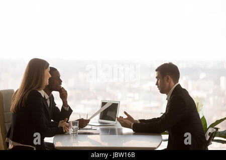 Friendly attentive hr managers interviewing vacancy applicant, multi-ethnic partners discussing new project idea sitting at office desk with cityscape Stock Photo