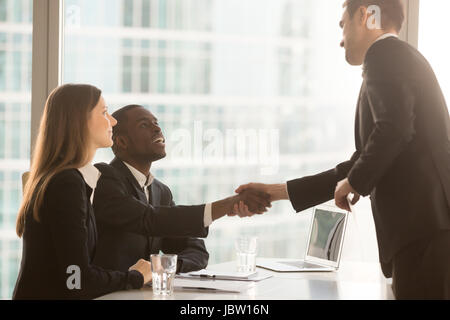 Friendly black and white recruiters sitting at office desk shaking hands with applicant just arrived for interview, diverse hr managers greeting candi Stock Photo