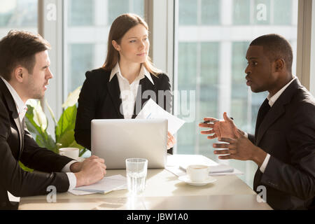 Multi-ethnic business team discussing project or idea sitting at office desk, partners working together, analyzing financial report, finding effective Stock Photo