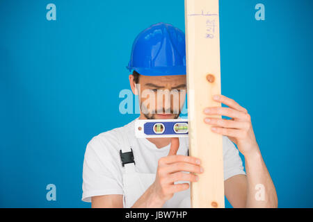 Handsome young workman in a hardhat using a spirit level holding it horizontally against a plank of wood against a blue background Stock Photo