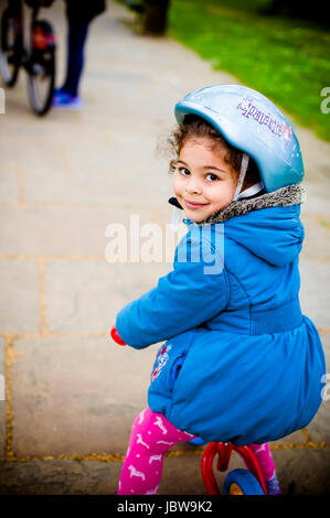 Four years old girl is riding her bike in the park. She is wearing a safety helmet and smiling to the camera. Stock Photo