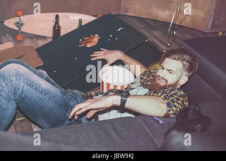 drunk bearded man lying on floor in messy room after party Stock Photo