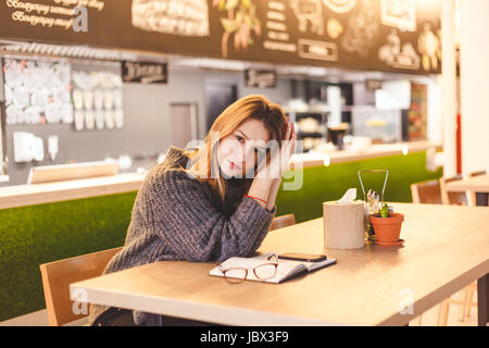 Young woman freelancer sitting in cafe after hard work day. Intelligent student rests in cafe after her lectures ends at University. Stock Photo