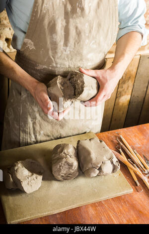 potter, clay, ceramics art concept - top view on hands of young master holding pieces of fireclay, male dressed in an apron, a ceramist with raw materials on table with sculpting tools set. Stock Photo