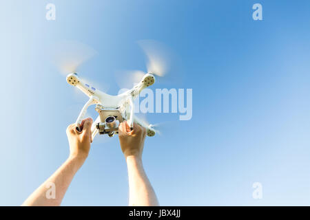 quadcopter outdoors, aerial imagery and recreation concept - closeup on human hands grip on frame of white quadrocopter flying on background of blue cloudless sky, male man caught flying drone. Stock Photo