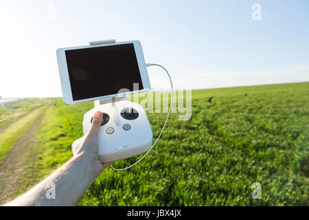 quadcopter flights outdoors, aerial imagery and tech hobby, recreation concept - white drone remote control in the man's hand of pilot on the background of green grass field and blue sky. Stock Photo