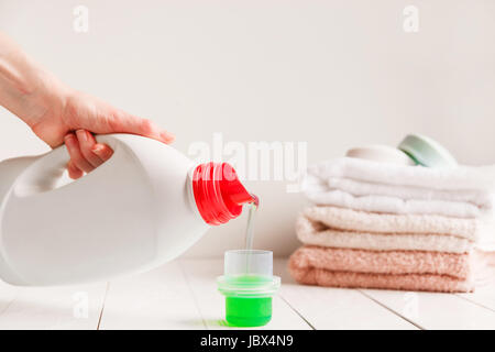 Close up of female hands pouring liquid laundry detergent into cap on white rustic table with towels on background in bathroom Stock Photo