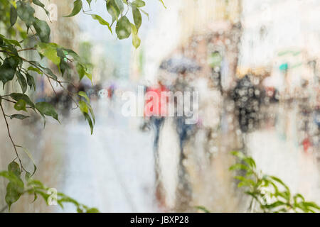Raindrops on window glass, few unrecognizable people rush home under umbrellas, abstract blurred background. Concept of seasons, weather, modern city. Stock Photo