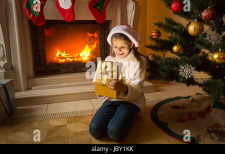 Cute girl sitting on floor at fireplace and receiving Christmas gift in golden box Stock Photo