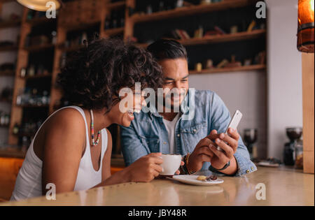 Happy young couple sitting at coffee shop looking at mobile phone and smiling. Young man and woman at cafe using smart phone. Stock Photo
