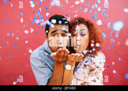 Close up of  young romantic couple blowing confetti in the air. Young man and woman blowing colorful star confetti out of their hand against red backg Stock Photo