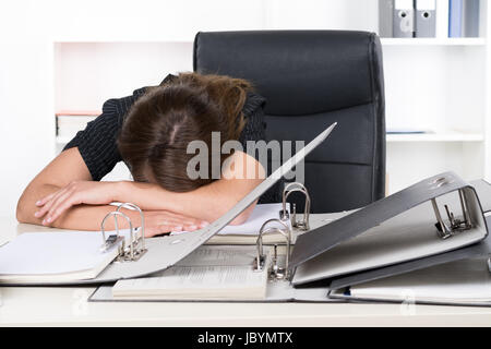 Eine junge, frustrierte Frau liegt mit ihrem Oberkörper vor einem Haufen von Akten auf Schreibtisch im Büro. Im Hintergrund steht ein Regal.