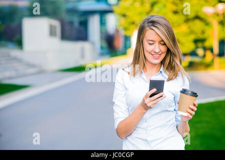 Young smart professional woman reading using phone. Female businesswoman reading news or texting sms on smartphone while drinking coffee on break from Stock Photo
