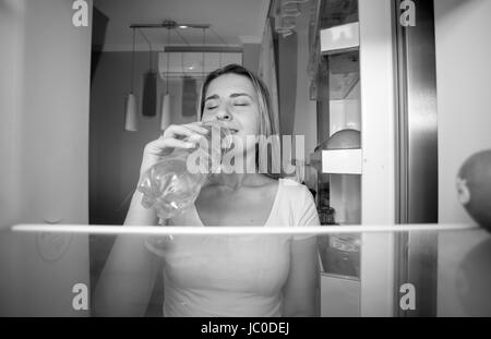 Black and white portrait of beautiful smiling woman taking water from fridge and drinking it. View from inside of open refrigerator Stock Photo