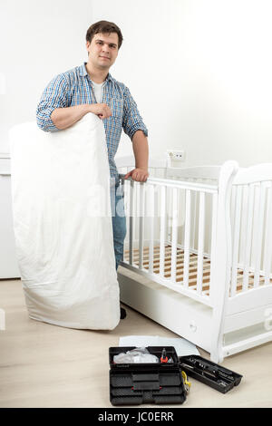 Young smiling man standing with mattress at disassembled baby's cot Stock Photo