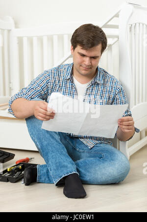 Young man reading manual on assembling wooden baby's crib Stock Photo