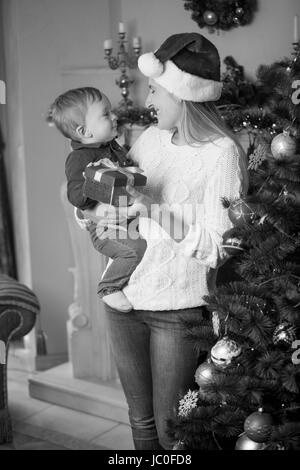 Black and white portrait of happy young mother in Santa hat and her 10 months old baby son posing at Christmas tree in living room Stock Photo