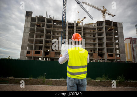 Rear view shot of engineer in hardhat and safety vest reading blueprints at building site Stock Photo