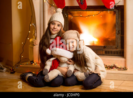 Happy smiling girls in sweater and hats warming up at fireplace at Christmas Stock Photo