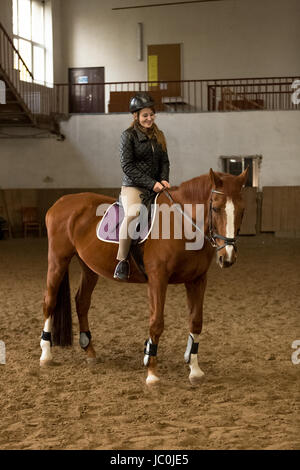 Beautiful young woman riding brown horse in indoor manege Stock Photo